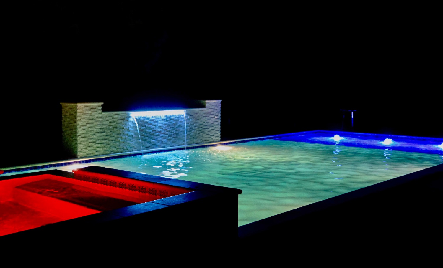Finished pool with blue lighting and a red-lit spa at night